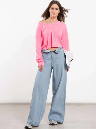 Eine junge Frau trägt das Outryght Kushi neon pink Oberteil, locker sitzend und schulterfrei, kombiniert mit weit geschnittenen Jeans und klobigen Sneakers. Sie hält eine weiße Baseballkappe in der Hand.