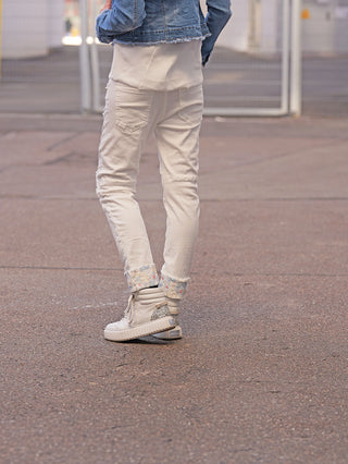 Person trägt OFF#DLY Denim White Flowers mit floralen Einsätzen an Knie und Saum, off-white, umgeschlagene Bündchen zeigen gestricktes Futter, kombiniert mit klobigen, glitzernden Sneakers auf einem gepflasterten Gehweg.