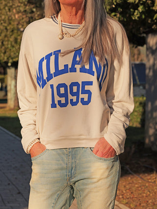 Person trägt OFF#DLY Sweat Milano stone, einen leichten Sweater mit „MILANO 1995“-Print, kombiniert mit Jeans und Perlenaccessoires. Perfekt für lässige Alltagslooks.