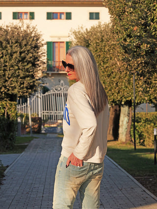 Frau in OFF#DLY Sweat Milano stone mit MILANO 1995 Print, leichtem Schnitt, stehend auf Gartenweg vor Villa. Kombiniert mit Jeans, trägt Sonnenbrille, Haare im Sonnenlicht.