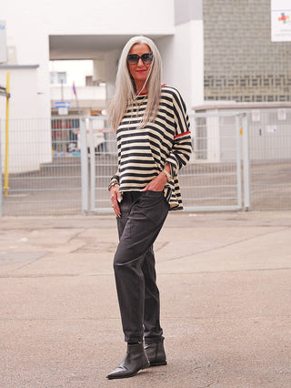 Die schlanke Frau trägt das OFF#DLY Ringel Shirt Ulla black/red, ein locker geschnittenes, langärmliges Streifenshirt mit modernen Kontrastdetails, kombiniert mit schwarzen Hosen und spitzen Stiefeletten.