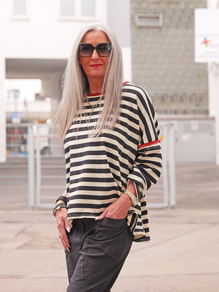 Person mit langen grauen Haaren trägt das OFF#DLY Ringel Shirt Ulla black/red mit schwarzen Hosen. Lässiges, gestreiftes Longsleeve mit roten Akzenten und entspannter Passform, ideal für Casual-Outfits.