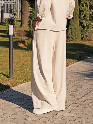 Person trägt die OFF#DLY Pant Lou eisgrau, eine weite, fließende Hose mit elastischem Bund und seitlichen Taschen, kombiniert mit weißen Slip-On-Sneakern und einer goldfarbenen Uhr.