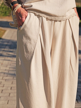 Nahaufnahme der OFF#DLY Pant Lou eisgrau: Weit geschnittene, cremefarbene Hose mit seitlichen Taschen, elastischem Bund und feiner Spitzenverzierung an den Kanten. Hände in den Taschen, Accessoires an den Handgelenken.