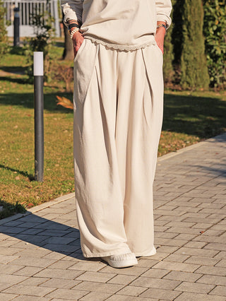 Person trägt OFF#DLY Pant Lou eisgrau, eine locker sitzende, weite Hose mit Seitentaschen. Kombiniert mit weißen Slippern auf einem Gehweg. Ideal für entspannte Eleganz und Komfort.