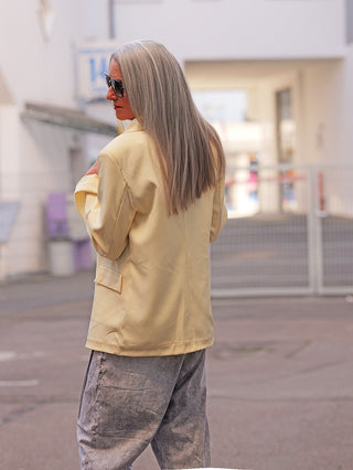 Person in gelber OFF#DLY Blazer Luise butteryellow, steht seitlich, trägt graue Hose und Sonnenbrille in städtischer Umgebung. Eleganter, lässiger Look mit weichem, fließendem Stoff.