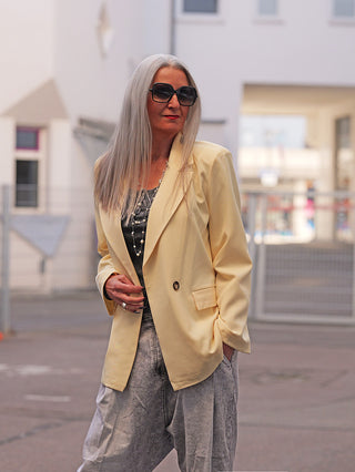 Ältere Frau trägt den OFF#DLY Blazer Luise butteryellow, kombiniert mit einer dunklen Oberbekleidung, einer langen Halskette und grauen Hosen, in städtischer Umgebung.