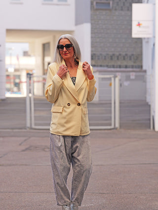 Eine Frau trägt den OFF#DLY Blazer Luise butteryellow, einen weich fallenden, doppelt geknöpften Blazer mit Reverskragen, ideal für lässige Eleganz. Sie hält die Blazer-Lapels in einer stilvollen Pose.