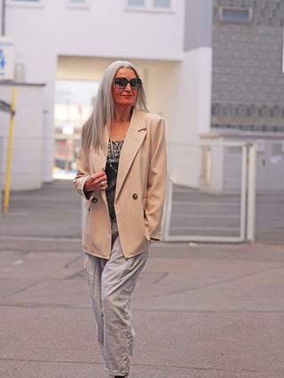 Frau im urbanen Umfeld trägt den OFF#DLY Blazer Luise beige, einen zart fallenden, doppelreihigen Blazer mit klassischem Reverskragen und aufgesetzten Taschen, kombiniert mit lockeren grauen Hosen.