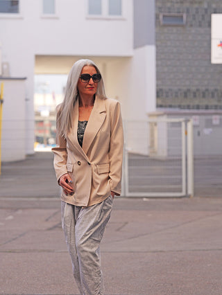 Frau in urbanem Hof trägt den OFF#DLY Blazer Luise beige, einen locker sitzenden, doppelt geknöpften Blazer, der moderne Lässigkeit und Eleganz ausstrahlt. Perfekt kombiniert mit grauer Hose.