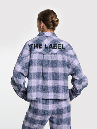 Person von hinten in ALIX Checked Blouse night blue mit Karomuster und Strass-Schriftzug „THE LABEL“ auf der Rückseite, kombiniert mit passenden Hosen, große Creolen sichtbar.