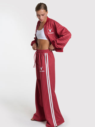 Model trägt ALIX Pinstripe Trainingpant Cerise, eine weite Hose mit Nadelstreifenmuster, weißen Seitenstreifen und Logo auf der Hüfte, kombiniert mit passender roter Bomberjacke und weißem Crop-Top.