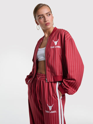 Junge Frau in ALIX Pinstripe Trainigns Jacket cerise, rotes Nadelstreifen-Design mit Bull-Stickerei und „THE LABEL“-Schriftzug, kombiniert mit passenden Trackpants und weißem Crop-Top.
