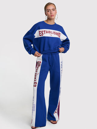 Frau in royalblauer ALIX Logo Tape Pant, weite Sweatpants mit weißen Seitenstreifen und rotem Schriftzug „ESTABLISHED ’14“, kombiniert mit passendem Sweatshirt und großen silbernen Ohrringen.