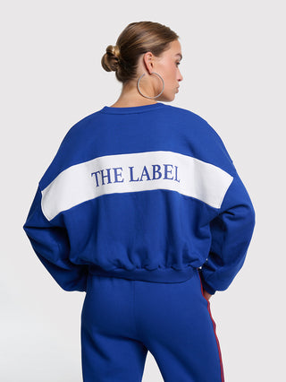 Frau von hinten in ALIX Logo Tape Sweater royal blue mit weißem Rückenband THE LABEL, silbernen Creolen, passender Hose mit Seitentrim. Sportlich-modernes Design mit lässiger Silhouette.