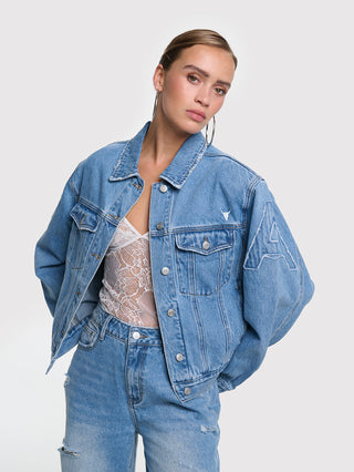 Eine junge Frau trägt die ALIX Soft Denim Patch Jacket blue mit auffälligen ALIX- und Bull-Patches auf den Ärmeln, kombiniert mit einer weißen Spitzenunterwäsche und hoch taillierten Jeans.