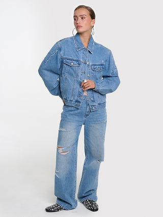 Modell trägt ALIX Soft Denim Patch Jacket blue mit kastigem Schnitt, Brusttaschen und Bull-Patches auf Ärmeln, kombiniert mit passenden Jeans und silberverzierten schwarzen Mules.
