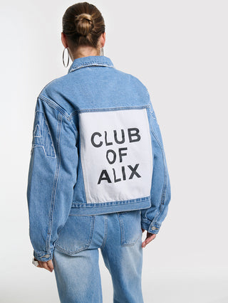 Rückansicht der ALIX Soft Denim Patch Jacket blue, mit großem weißen CLUB OF ALIX-Rückenpatch. Die Jacke zeigt charakteristische ALIX- und Bull-Patches auf den Ärmeln, getragen von Frau mit Dutt.