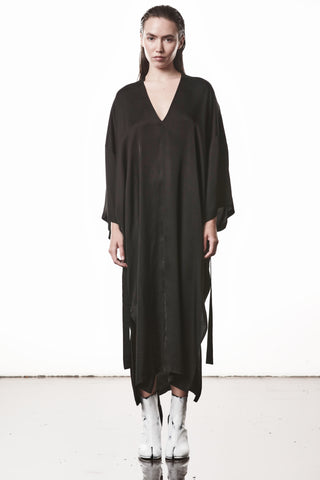 Die Thom Krom Dress WD 61 black zeigt ein oversized Sateen-Kleid mit V-Ausschnitt, asymmetrischem Saum und Kimono-Ärmeln, getragen von einem Modell mit weißen Stiefeln.