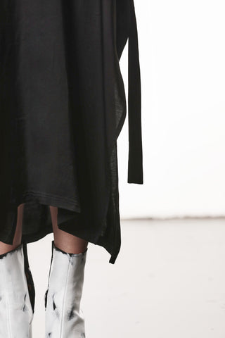 Person trägt Thom Krom Dress WD 61 black, ein lockeres, asymmetrisches, knielanges schwarzes Kleid. Kombiniert mit auffälligen, metallisch-silbernen Stiefeln. Kleid zeigt modernen Minimalismus und avantgardistische Eleganz.