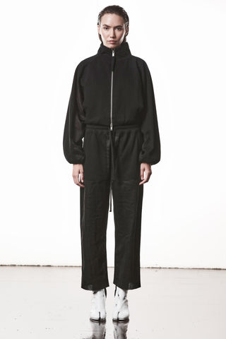 Frau trägt die Thom Krom Jacke WSJ 497 black mit leicht oversized Fit, hohem Stehkragen und Daumenöffnungen an den Ärmeln, kombiniert mit passenden weiten Hosen und weißen Split-Toe-Stiefeletten.