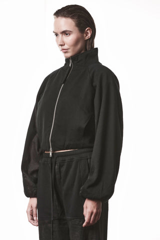 Eine junge Frau trägt die Thom Krom Jacke WSJ 497 black mit hohem Kragen und Reißverschluss, kombiniert mit weiten schwarzen Hosen; die Jacke hat markante, strukturierte, leicht oversized Ärmel.