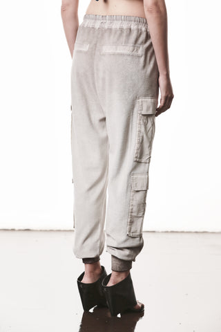 Die Thom Krom Hose WST 427 stone zeigt einen urbanen Avantgarde-Look mit Cargo-Taschen, elastischem Tunnelzugbund und schmal zulaufendem Bein, getragen mit schwarzen, offenen Keilsandalen.