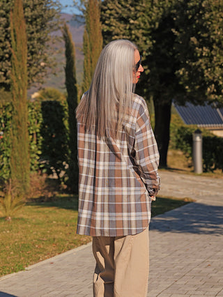 Eine Person trägt die OFF#DLY Bluse Susi blue/brown mit Oversize-Schnitt und Karomuster, stehend auf einem Weg in einem Garten, umgeben von gepflegter Landschaft.