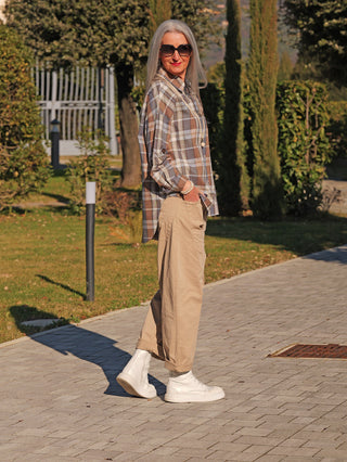 Ältere Person trägt die OFF#DLY Bluse Susi blue/brown in lockerem Oversize-Schnitt und Karomuster, kombiniert mit beigen Hosen und weißen Sneakers, stehend in einem parkähnlichen Umfeld.