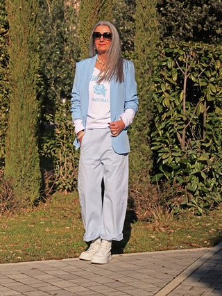Frau trägt den OFF#DLY Blazer Diana lightblue, offen über weißem T-Shirt mit blauer Rose, kombiniert mit passenden weiten Hosen und weißen Sneakers, in sonniger Parkumgebung.