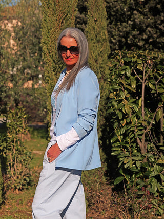 Person trägt den OFF#DLY Blazer Diana lightblue, kombiniert mit einer weißen Top und passenden hellblauen Hose, während sie in einem sonnigen Garten mit Perlenkette und dunkler Sonnenbrille steht.