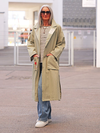Ältere Frau trägt den OFF#DLY Mantel Jessy Verde: khakifarbener Trenchcoat mit großen Taschen, darunter gestreiftes Oberteil, blaue Jeans und weiße Sneaker, Hände in Manteltaschen, Namensschild an der Lapel.