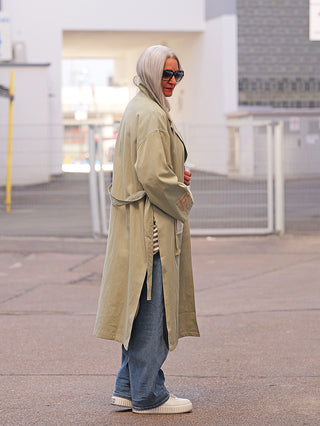 Person trägt den OFF#DLY Mantel Jessy Verde, ein lässiger, knie-langer Trenchcoat mit markanten Details. Kombiniert mit Streifen-Top, blauen Jeans und weißen Sneakers für einen urbanen Look.