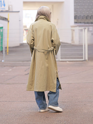 Person in OFF#DLY Mantel Jessy Verde, knielang mit Gürtel, trägt locker sitzend über blauen Jeans. Im Hintergrund ein Zaun und ein Gebäude.