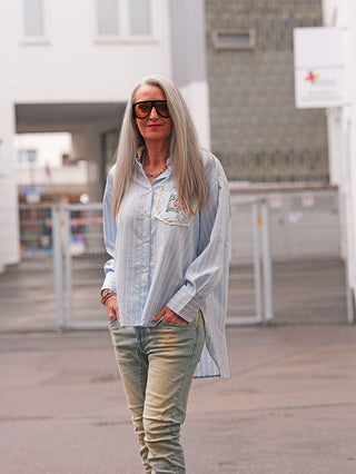 Ältere Person im Freien trägt die OFF#DLY Bluse Ancona stripes lightblue, eine oversized, gestreifte Hemdbluse mit Brusttaschen-Detail, kombiniert mit Jeans, Sonnenbrille und Schmuck, betont urban-lässigen Stil.