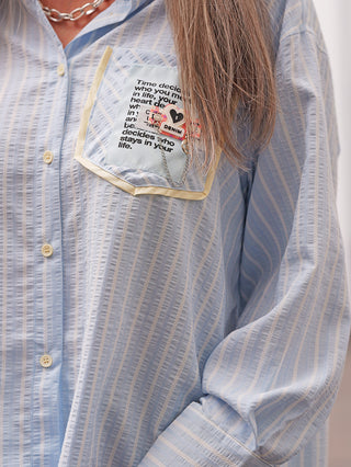 Nahaufnahme des Torsos mit der OFF#DLY Bluse Ancona stripes lightblue, einem Oversize-Hemd mit hellblauen Streifen, gelb umrandeter Brusttasche mit Motivationszitat und dekorativen Metallabzeichen.