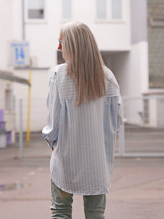 Person von hinten mit grauem Haar trägt die OFF#DLY Bluse Ancona stripes lightblue, eine locker sitzende, hellblaue Streifenbluse mit Oversize-Schnitt, auf einem gepflasterten Platz vor einem weißen Gebäude.