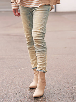 Person trägt OFF#DLY Denim Genua midblue: Leicht verwaschene, eng anliegende Jeans mit dezenten Used-Details und umgeschlagenen Säumen, kombiniert mit beigefarbenen, spitzen Ankle Boots.