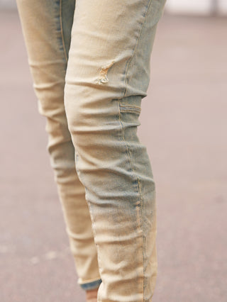 Nahaufnahme von Beinen in OFF#DLY Denim Genua midblue Jeans, schmal geschnitten mit kleinem Riss über dem Knie, auf gepflastertem Untergrund stehend, betont durch dezenten Used-Look und Stretch-Komfort.