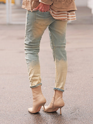 Person trägt OFF#DLY Denim Genua midblue, eine schlanke, zweifarbige Jeans mit umgeschlagenen Knöcheln, Hand in der Gesäßtasche, kombiniert mit spitzen Stiefeletten auf nassem Pflaster.