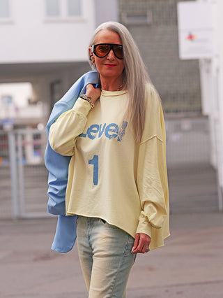 Ältere Frau trägt das OFF#DLY Sweat Level 1 yellow, ein locker geschnittenes, leichtes Sweatshirt mit Level 1 Print, kombiniert mit einer hellblauen Jacke und Jeans in urbaner Umgebung.