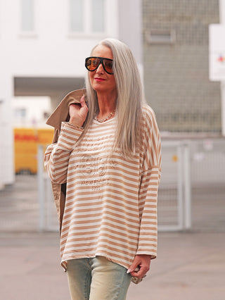 Eine reife Frau trägt das OFF#DLY Longsleeve Lousiana beige/white mit Streifen, kombiniert mit hellen Jeans, Sonnenbrille und Schmuck, in einem urbanen Außenbereich, was einen lässig-eleganten Look zeigt.