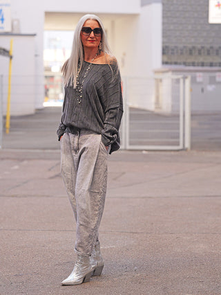 Frau mit grauen Haaren trägt OFF#DLY Pulli Agathe black, ein locker gestrickter Pullover mit Strukturmuster und ausgefranstem Saum, kombiniert mit Jeans und silbernen Stiefeletten in urbaner Umgebung.