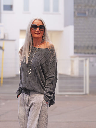 Person trägt OFF#DLY Pulli Agathe black mit vertikalem Strukturmuster und lockerem Schnitt, der über eine Schulter fällt, kombiniert mit locker sitzenden Jeans und einer langen, perlenbesetzten Halskette.