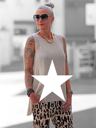 Dame mit silbernem Haar trägt das ärmellose OFF#DLY Top Taranto white in Beige, verziert mit einem großen weißen Stern, kombiniert mit hoch taillierten Leopardenmuster-Hosen, Schmuck und Tattoos.
