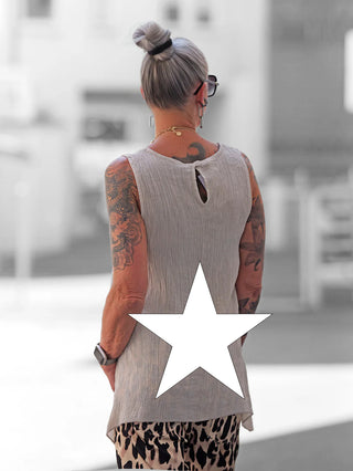 Rückansicht einer Frau in urbanem Umfeld, die das ärmellose OFF#DLY Top Taranto trägt, mit Crinkle-Struktur und verlängertem Rückenteil, passend bis Größe 44. Tätowierte Arme und silberne Haare im Dutt.