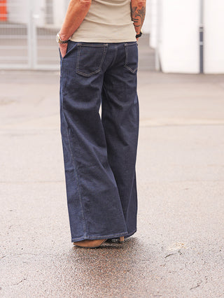 Eine Person trägt True Religion Lauren RAW Jeans mit weitem Bein, dunklem Raw-Wash, in die Gesäßtaschen gesteckte Hände, sichtbar mit beigem ärmellosen Top und tätowiertem Unterarm.