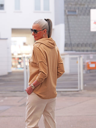 Die Frau trägt das OFF#DLY LS Thea camel Hoodie mit großem Herzmotiv, das sportlichen Komfort und Eleganz vereint, perfekt kombiniert mit hellen Beige-Tönen für einen stilvollen Look.