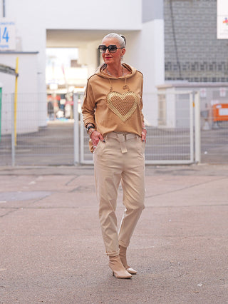 Eine Frau trägt den OFF#DLY LS Thea camel Hoodie mit einem goldenen Herzmotiv, kombiniert mit beigefarbenen, taillierten Hosen und passenden Stiefeletten, in einer urbanen Umgebung.