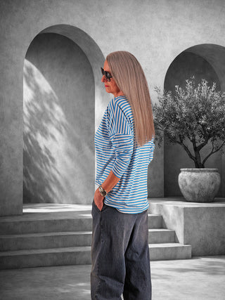 Person im Profil trägt das OFF#DLY Ringelshirt Frida türkis/white, ein locker geschnittenes, gestreiftes Longsleeve, kombiniert mit dunkler Hose vor minimalistischer Kulisse mit architektonischen Elementen und einem großen Pflanzkübel.